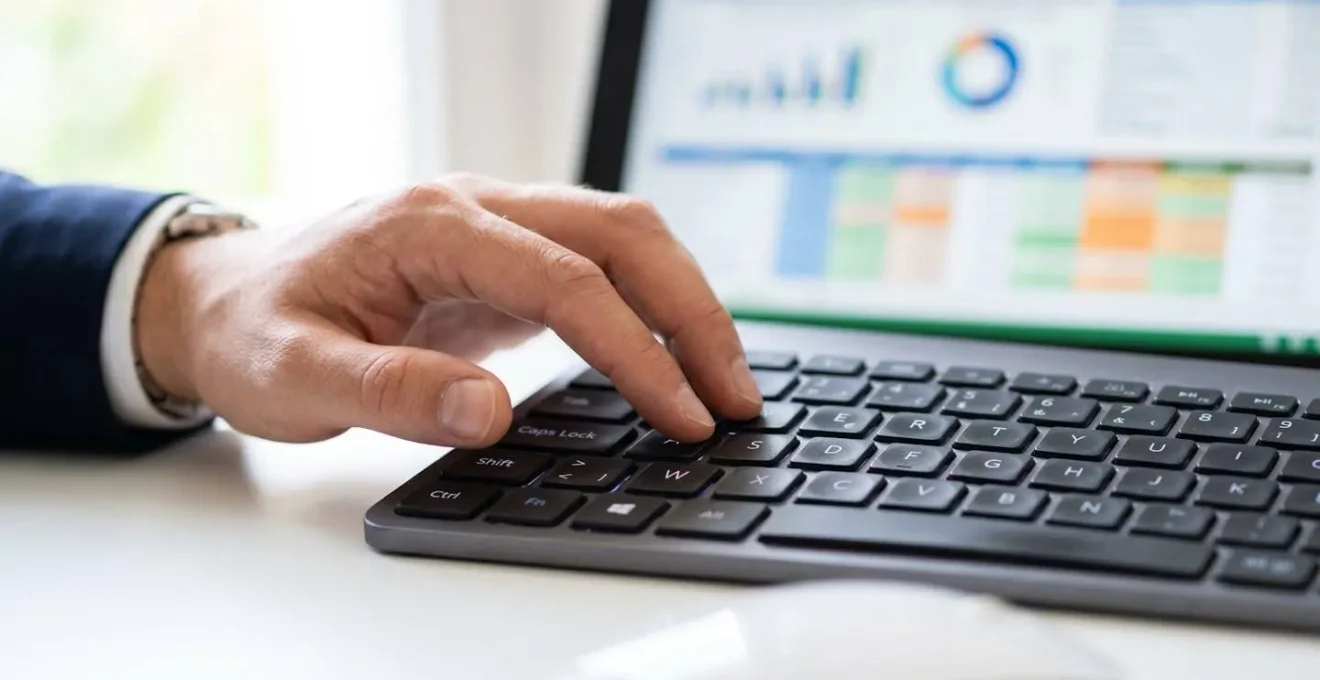 Gros plan sur une main tapant sur un clavier d'ordinateur portable avec un tableur financier visible mais totalement flouté en arrière-plan